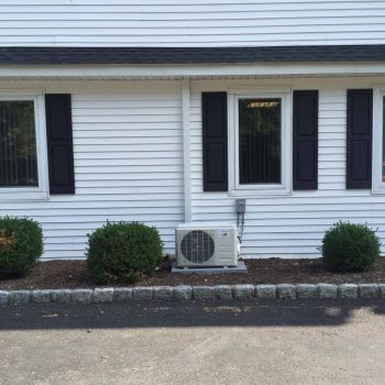 Outdoor air conditioning unit installed beside a white house with windows and shrubs